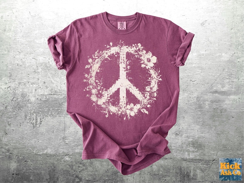 May include: A short-sleeved crew neck t-shirt in a muted berry-purple color. The center of the shirt features a large, off-white graphic of a peace sign surrounded by a detailed floral wreath pattern. The fabric has a slightly weathered texture.