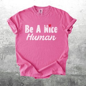 K&ouml;nnte beinhalten: Ein pinkfarbenes T-Shirt mit dem wei&szlig;en Schriftzug "Be A Nice Human" und einem kleinen roten Herz &uuml;ber dem Wort "Nice". Das T-Shirt ist gefaltet und hat hochgekrempelte &Auml;rmel. Der Hintergrund ist eine strukturierte, graue Oberfl&auml;che.