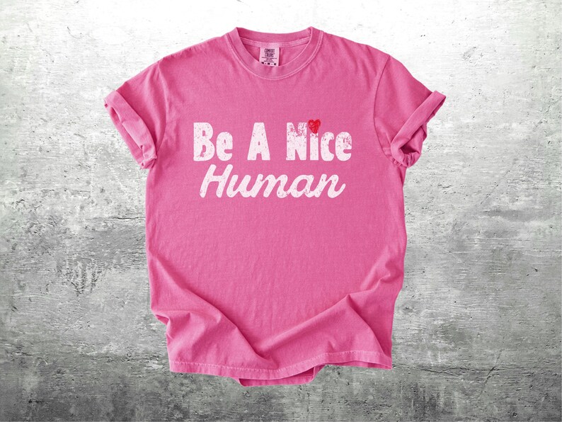 K&ouml;nnte beinhalten: Ein pinkfarbenes T-Shirt mit dem wei&szlig;en Schriftzug "Be A Nice Human" und einem kleinen roten Herz &uuml;ber dem Wort "Nice". Das T-Shirt ist gefaltet und hat hochgekrempelte &Auml;rmel. Der Hintergrund ist eine strukturierte, graue Oberfl&auml;che.
