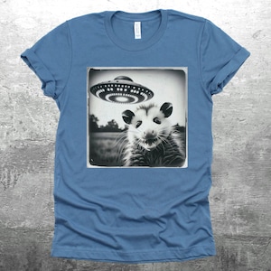 May include: A blue t-shirt with a black and white image of an opossum looking up at a flying saucer.