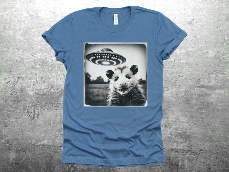 May include: A blue t-shirt with a black and white image of an opossum looking up at a flying saucer.