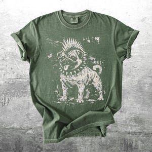 May include: Olive green t-shirt featuring a distressed graphic of a pug with a mohawk and studded collar. The dog's tongue is sticking out. The shirt has rolled-up sleeves and a crew neck.
