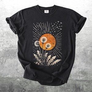 May include: Black t-shirt featuring a dandelion design. The graphic includes an orange sun, white rays, and dandelion seed heads. The shirt has rolled-up sleeves and a crew neck.