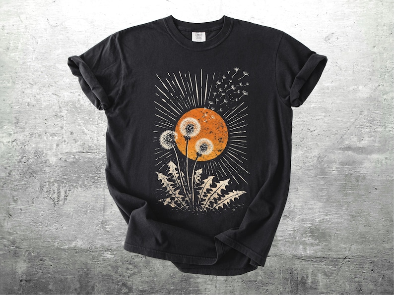 May include: Black t-shirt featuring a dandelion design. The graphic includes an orange sun, white rays, and dandelion seed heads. The shirt has rolled-up sleeves and a crew neck.