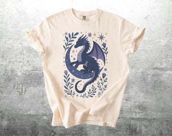 May include: Cream-colored t-shirt featuring a dark blue dragon perched on a crescent moon. The design includes purple and blue floral accents and stars. The shirt is made of a soft material.
