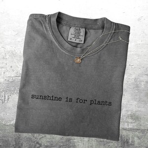 May include: A grey Comfort Colors t-shirt with the text "sunshine is for plants" printed in black. A silver necklace with a small pendant rests on the shirt. The shirt is folded on a grey surface.