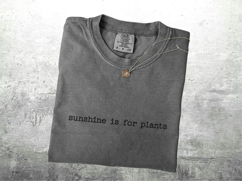 May include: A grey Comfort Colors t-shirt with the text "sunshine is for plants" printed in black. A silver necklace with a small pendant rests on the shirt. The shirt is folded on a grey surface.