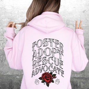 Pu&ograve; includere: Felpa con cappuccio rosa con il testo "FOSTER ADOPT RESCUE ADVOCATE" in lettere nere sul retro. Un rosa rossa con foglie verdi e due impronte di zampa nere sono sotto il testo.