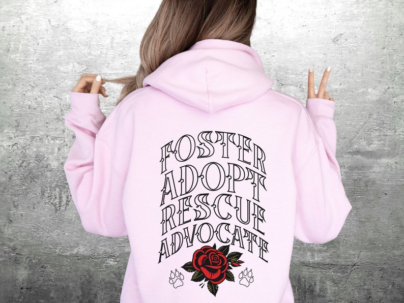 Pu&ograve; includere: Felpa con cappuccio rosa con il testo "FOSTER ADOPT RESCUE ADVOCATE" in lettere nere sul retro. Un rosa rossa con foglie verdi e due impronte di zampa nere sono sotto il testo.