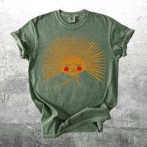 K&ouml;nnte beinhalten: Gr&uuml;nes T-Shirt mit einem Sonnengrafik. Die Sonne hat orangefarbene Strahlen, ein l&auml;chelndes Gesicht und rote Wangen &uuml;ber einer Bergkette. Das T-Shirt hat einen Vintage-Look und hochgekrempelte &Auml;rmel. Das Design ist mittig auf der Vorderseite.