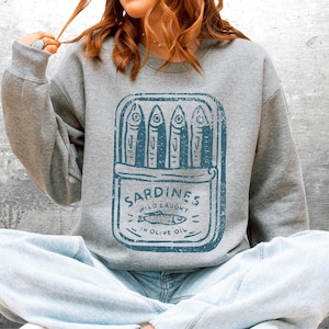May include: A gray sweatshirt featuring a blue graphic of a sardine can with the text "Sardines Wild Caught in Olive Oil." The design includes stylized fish illustrations. The sweatshirt is worn by a person.