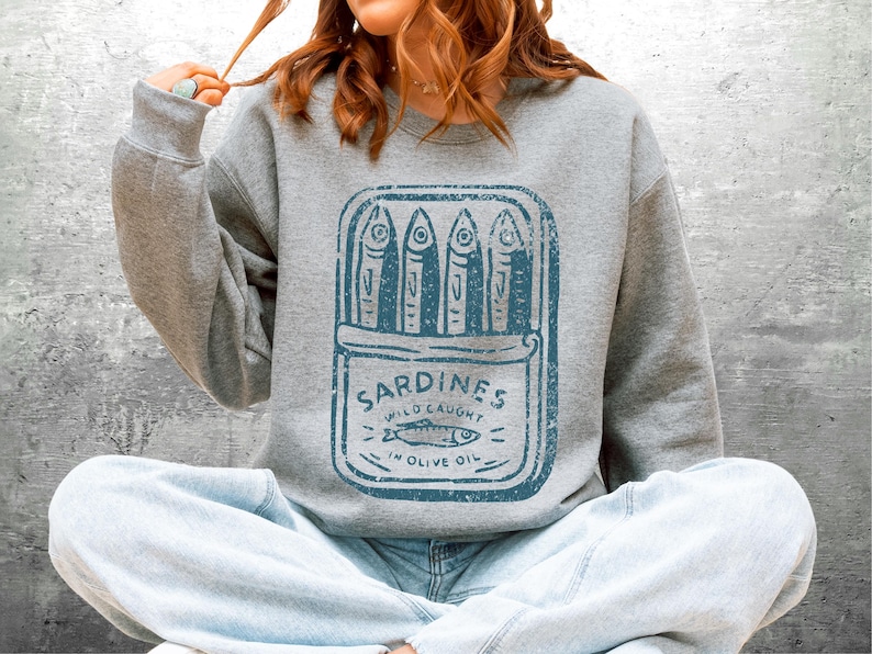 May include: A gray sweatshirt featuring a blue graphic of a sardine can with the text "Sardines Wild Caught in Olive Oil." The design includes stylized fish illustrations. The sweatshirt is worn by a person.