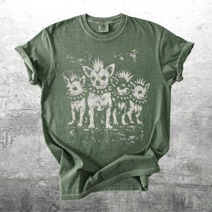 May include: Olive green t-shirt featuring a distressed graphic of four stylized chihuahua dogs with mohawks and studded collars. The dogs are standing in a row. The shirt has a crew neck and short sleeves.