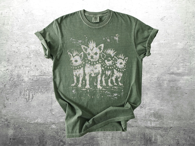May include: Olive green t-shirt featuring a distressed graphic of four stylized chihuahua dogs with mohawks and studded collars. The dogs are standing in a row. The shirt has a crew neck and short sleeves.