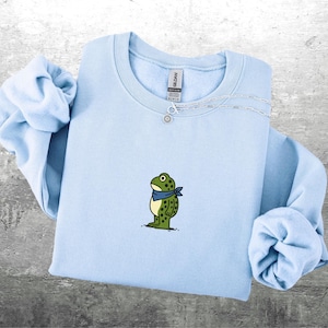 Puede incluir: Una sudadera azul claro con una rana de dibujos animados con una bandana azul. La rana es verde con manchas negras. Un collar plateado con un peque&ntilde;o colgante est&aacute; en la sudadera. Las mangas est&aacute;n dobladas.