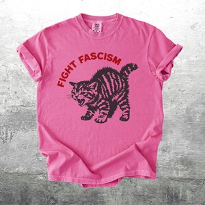 K&ouml;nnte beinhalten: Rosafarbenes T-Shirt mit dem roten Schriftzug "FIGHT FASCISM", der sich &uuml;ber einer schwarz-braun gestreiften Katzenzeichnung w&ouml;lbt. Die Katze ist mit offenem Maul dargestellt. Das T-Shirt liegt auf einer grauen Oberfl&auml;che.