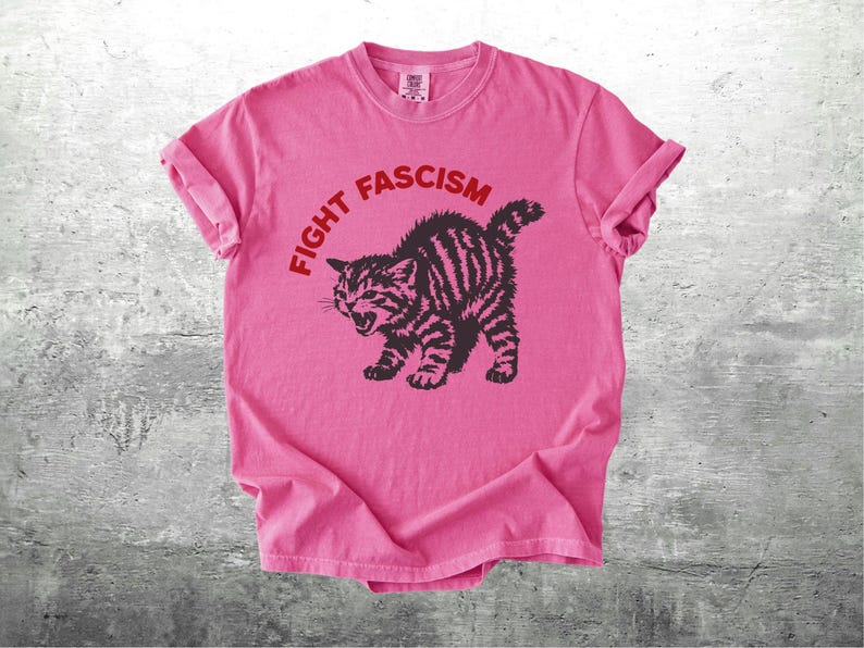 K&ouml;nnte beinhalten: Rosafarbenes T-Shirt mit dem roten Schriftzug "FIGHT FASCISM", der sich &uuml;ber einer schwarz-braun gestreiften Katzenzeichnung w&ouml;lbt. Die Katze ist mit offenem Maul dargestellt. Das T-Shirt liegt auf einer grauen Oberfl&auml;che.