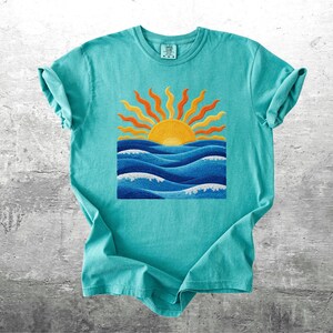 May include: A turquoise t-shirt featuring a vibrant graphic of a yellow sun with orange rays above blue ocean waves. The design is centered on the shirt, creating a summery and cheerful aesthetic. The shirt has short sleeves and a crew neck.