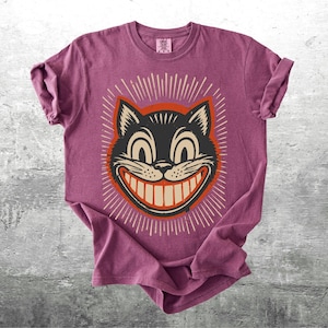 Op de afbeelding: Een mauve t-shirt met korte mouwen en een afbeelding van een zwarte kattenkop met een brede, tandige grijns. De kat is omlijnd in oranje en omringd door witte stralende lijnen. Het shirt heeft een ronde hals en een label in de kraag.
