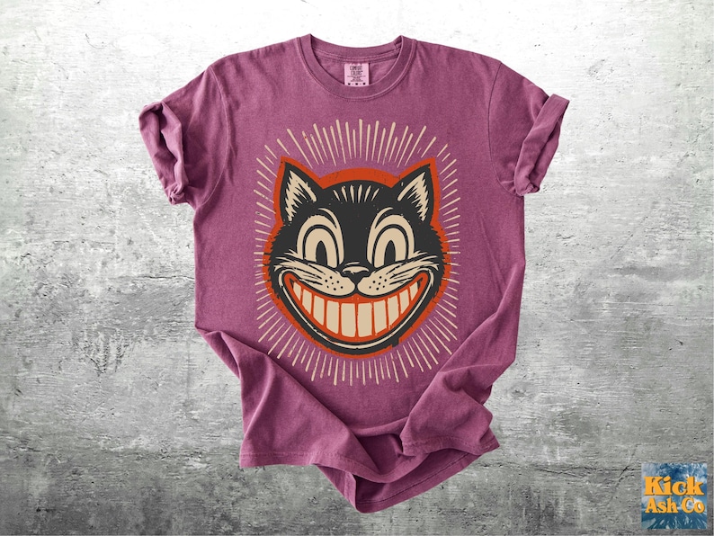 Op de afbeelding: Een mauve t-shirt met korte mouwen en een afbeelding van een zwarte kattenkop met een brede, tandige grijns. De kat is omlijnd in oranje en omringd door witte stralende lijnen. Het shirt heeft een ronde hals en een label in de kraag.