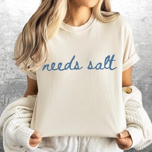 May include: A white t-shirt with the text "needs salt" written in blue cursive.