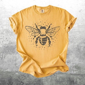 May include: Mustard yellow t-shirt featuring a detailed black illustration of a bee with outstretched wings and surrounding stars. The shirt has rolled-up sleeves and a crew neck, and is made of a soft, comfortable material.