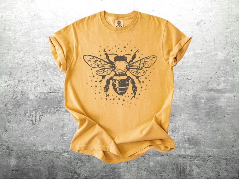 May include: Mustard yellow t-shirt featuring a detailed black illustration of a bee with outstretched wings and surrounding stars. The shirt has rolled-up sleeves and a crew neck, and is made of a soft, comfortable material.