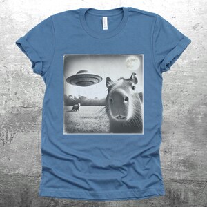 K&ouml;nnte beinhalten: Ein blaues T-Shirt mit einem Schwarzwei&szlig;bild eines Capybaras, das zu einem fliegenden Untertasse aufblickt. Das Bild ist im Vintage-Stil mit einem Mond im Hintergrund.