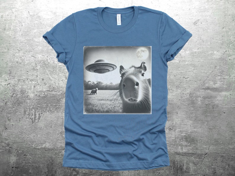 K&ouml;nnte beinhalten: Ein blaues T-Shirt mit einem Schwarzwei&szlig;bild eines Capybaras, das zu einem fliegenden Untertasse aufblickt. Das Bild ist im Vintage-Stil mit einem Mond im Hintergrund.