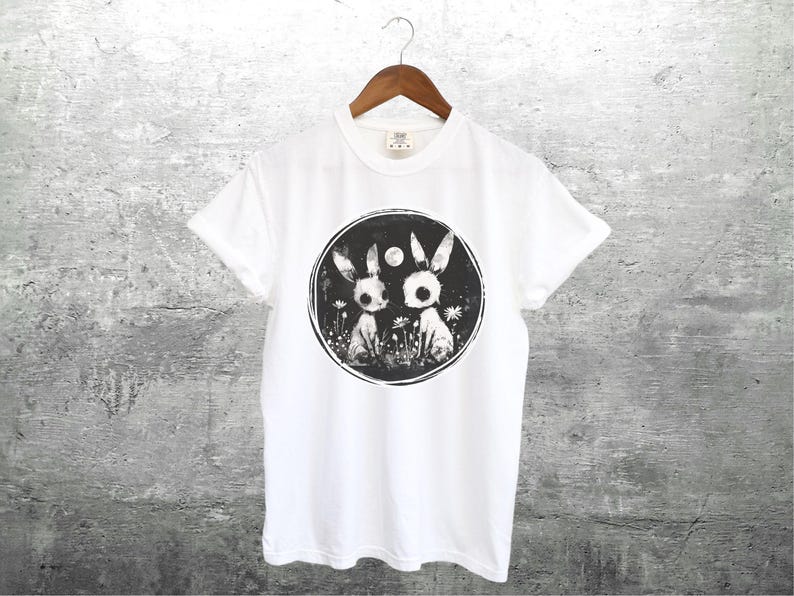 Op de afbeelding: Wit T-shirt met een zwart-witte grafische print van twee konijnen in een cirkel met een maan en sterren. De konijnen zijn gestileerd en hebben grote ogen.