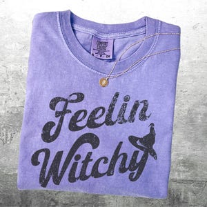K&ouml;nnte beinhalten: Ein fliederfarbenes Comfort Colors T-Shirt mit dem Schriftzug "Feelin Witchy" in verblichenen schwarzen Buchstaben. Eine goldene Halskette mit einem kleinen Anh&auml;nger liegt auf dem Shirt. Das Shirt liegt auf einer grauen Oberfl&auml;che.