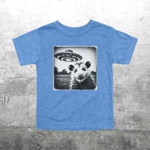 May include: A light blue toddler t-shirt with a black and white image of an opossum and a UFO. The opossum is looking at the UFO. The image is in a square frame.