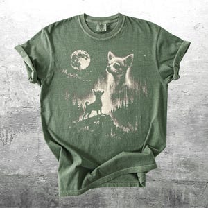 May include: Green t-shirt featuring a graphic of a Chihuahua dog under a full moon. The design includes a silhouette of the dog on a hill and a larger image of the dog's face. The shirt has a vintage, distressed look.