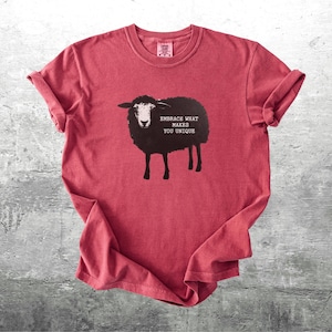 Puede incluir: Camiseta roja con un dise&ntilde;o de oveja negra y el texto "Embrace What Makes You Unique". La oveja tiene la cara blanca y est&aacute; sobre cuatro patas. Las mangas est&aacute;n enrolladas. La camiseta est&aacute; sobre una superficie gris.