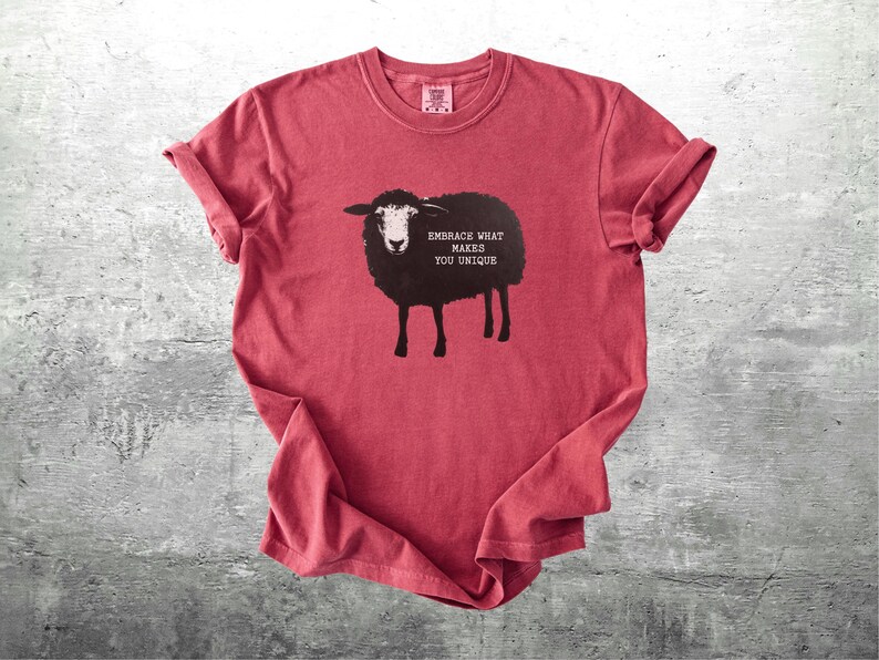 Puede incluir: Camiseta roja con un dise&ntilde;o de oveja negra y el texto "Embrace What Makes You Unique". La oveja tiene la cara blanca y est&aacute; sobre cuatro patas. Las mangas est&aacute;n enrolladas. La camiseta est&aacute; sobre una superficie gris.