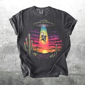 May include: A charcoal gray t-shirt featuring a graphic of a possum being abducted by a flying saucer in a desert landscape. The scene includes cacti, a sunset, and a vibrant beam of light. The text Kick Ash Co is visible in the bottom corner.