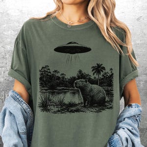 May include: Olive green t-shirt featuring a black and white graphic of a capybara near a body of water, with a UFO above. The shirt has rolled-up sleeves and a relaxed fit. The text "Kick Ash Co" is in the bottom right corner.