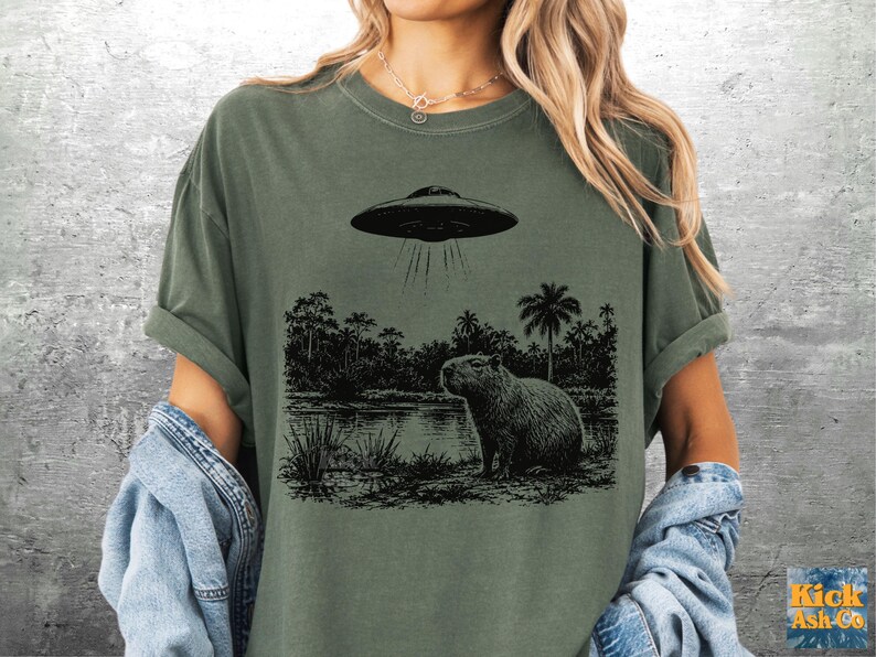 May include: Olive green t-shirt featuring a black and white graphic of a capybara near a body of water, with a UFO above. The shirt has rolled-up sleeves and a relaxed fit. The text "Kick Ash Co" is in the bottom right corner.