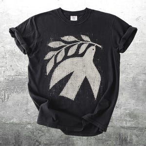 May include: A black short-sleeve t-shirt featuring a distressed, off-white graphic print of a dove carrying an olive branch in its beak. The design has a textured, weathered appearance centered on the chest of the garment, which has a crew neckline.