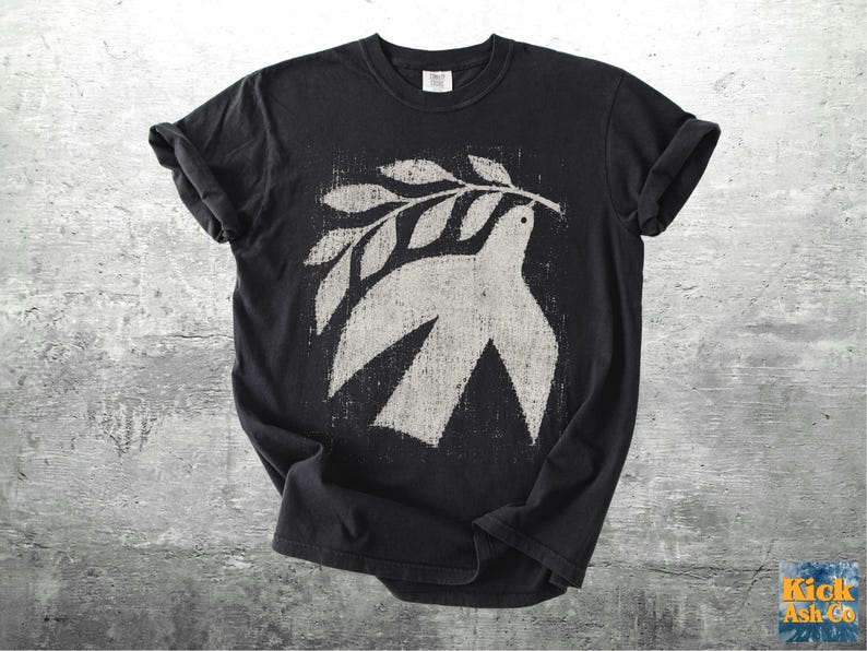 May include: A black short-sleeve t-shirt featuring a distressed, off-white graphic print of a dove carrying an olive branch in its beak. The design has a textured, weathered appearance centered on the chest of the garment, which has a crew neckline.