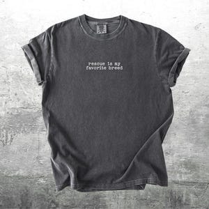 May include: Dark gray t-shirt with rolled-up sleeves. The shirt features the text "rescue is my favorite breed" in a simple, white font. The shirt is made of a soft material and is displayed on a textured gray background.
