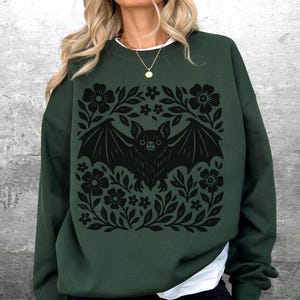 May include: Dark green sweatshirt featuring a black bat design surrounded by floral elements. The bat has outstretched wings and is centered within a square floral frame. The sweatshirt has long sleeves and a white collar.