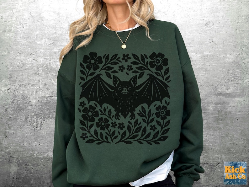 May include: Dark green sweatshirt featuring a black bat design surrounded by floral elements. The bat has outstretched wings and is centered within a square floral frame. The sweatshirt has long sleeves and a white collar.