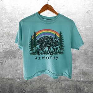 May include: A light blue short-sleeve t-shirt featuring a black graphic of a bear walking through a forest under a colorful rainbow. The text JIMOTHY is printed in black below the bear. The shirt is displayed on a wooden hanger.