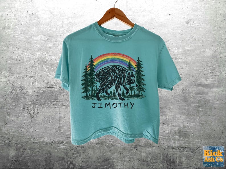 May include: A light blue short-sleeve t-shirt featuring a black graphic of a bear walking through a forest under a colorful rainbow. The text JIMOTHY is printed in black below the bear. The shirt is displayed on a wooden hanger.
