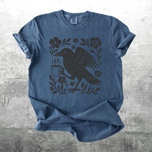 May include: Blue t-shirt with a black graphic of a crow holding a coffee cup, surrounded by flowers and leaves. The design is centered on the shirt. The shirt has short sleeves and a crew neck.