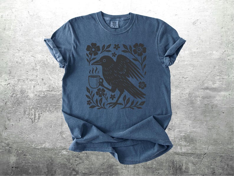 May include: Blue t-shirt with a black graphic of a crow holding a coffee cup, surrounded by flowers and leaves. The design is centered on the shirt. The shirt has short sleeves and a crew neck.