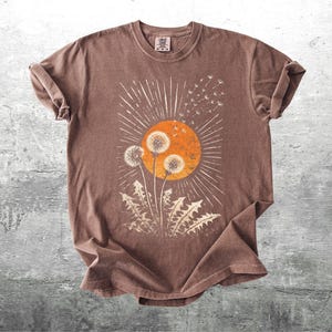 May include: A brown t-shirt featuring a dandelion design with an orange sun and white rays. The graphic includes dandelion seed heads and leaves. The shirt is made of a soft material and has rolled-up sleeves.