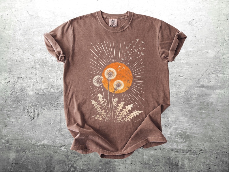 May include: A brown t-shirt featuring a dandelion design with an orange sun and white rays. The graphic includes dandelion seed heads and leaves. The shirt is made of a soft material and has rolled-up sleeves.