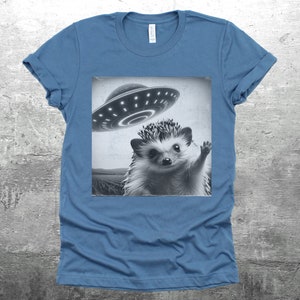 May include: A blue t-shirt with a black and white image of a hedgehog waving at a flying saucer.